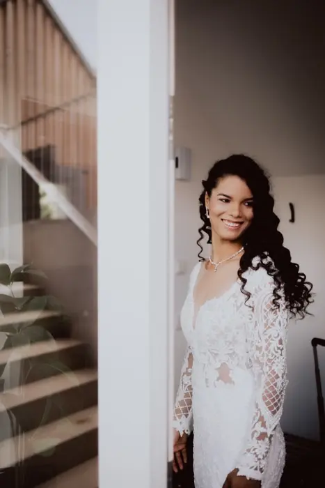 Hochzeitsfotograf Bayern Braut in Spitzenhochzeitskleid lächelt neben einer Tür Dokumentarische Editorial Hochzeitsfotografie