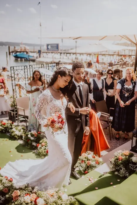 Hochzeitsfotograf Bayern Brautpaar während einer Hochzeitszeremonie am See mit Gästen und Blumendekoration Dokumentarische Editorial Hochzeitsfotografie