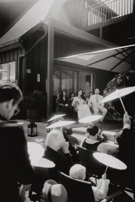 Hochzeitsfotograf Bayern Schwarzweiß-Hochzeitsfoto mit Braut, Bräutigam und Gästen mit Sonnenschirmen Dokumentarische Editorial Hochzeitsfotografie