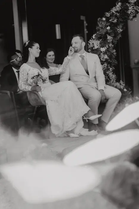 Hochzeitsfotograf Bayern Hochzeitspaar lachend auf Stühlen bei Trauung mit Blumendekoration Dokumentarische Editorial Hochzeitsfotografie