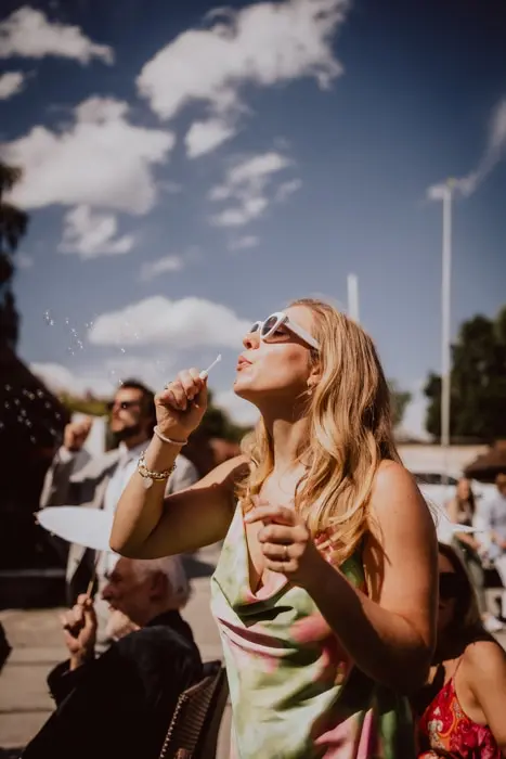 Hochzeitsfotograf Bayern Frau bei sonnigem Wetter, Sonnenbrille tragend, während sie Seifenblasen macht Dokumentarische Editorial Hochzeitsfotografie