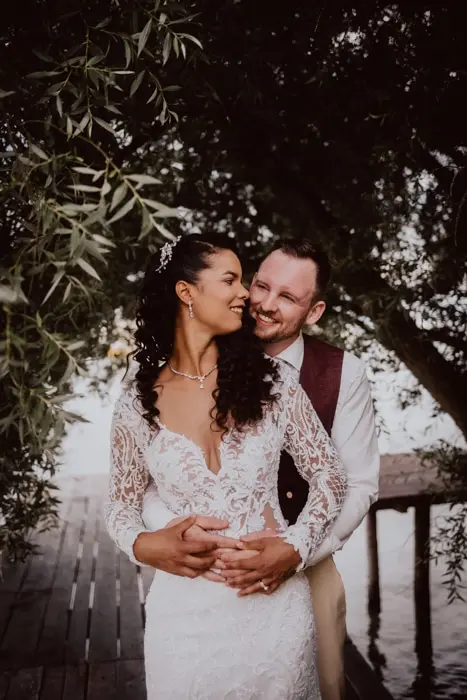 Hochzeitsfotograf Bayern Ein Brautpaar umarmt sich unter einem Baum am Wasser Dokumentarische Editorial Hochzeitsfotografie