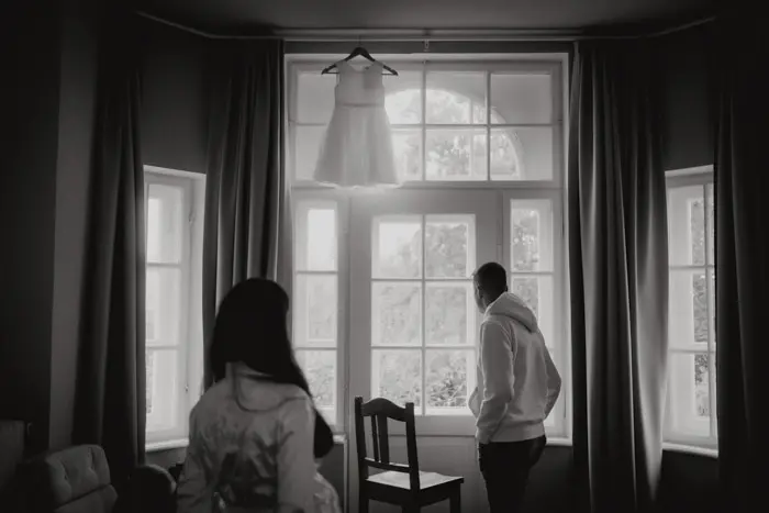 Hochzeitsfotograf Bayern Schwarzweiß-Foto mit zwei Personen und einem aufgehängten Kleid vor einem Fenster Dokumentarische Editorial Hochzeitsfotografie