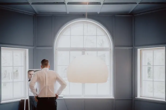 Hochzeitsfotograf Bayern Mann betrachtet sich im Spiegel in einem eleganten Raum mit großen Fenstern Dokumentarische Editorial Hochzeitsfotografie