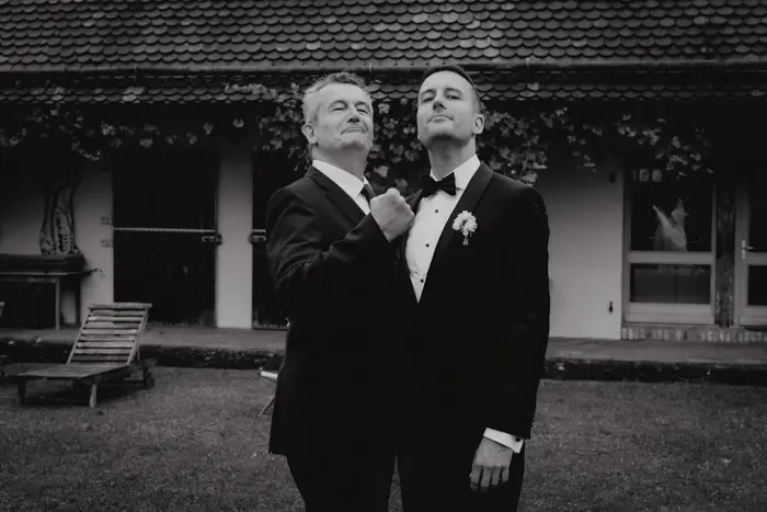 Hochzeitsfotograf Bayern Zwei Männer in Anzügen posieren selbstbewusst vor einem Haus Dokumentarische Editorial Hochzeitsfotografie