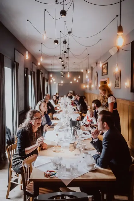 Hochzeitsfotograf Bayern Elegantes Restaurant mit Gästen an langen Holztischen und dezenter Beleuchtung Dokumentarische Editorial Hochzeitsfotografie