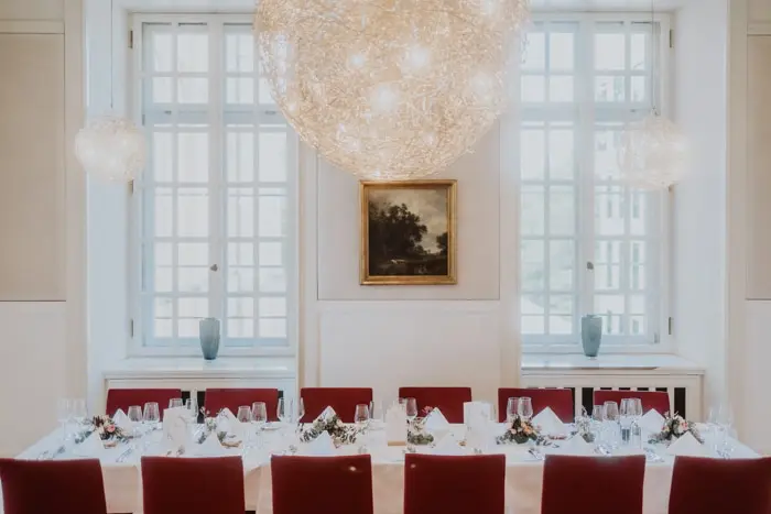 Hochzeitsfotograf Bayern Eleganter Speisesaal mit rot gepolsterten Stühlen und großer Kunstlampe Dokumentarische Editorial Hochzeitsfotografie