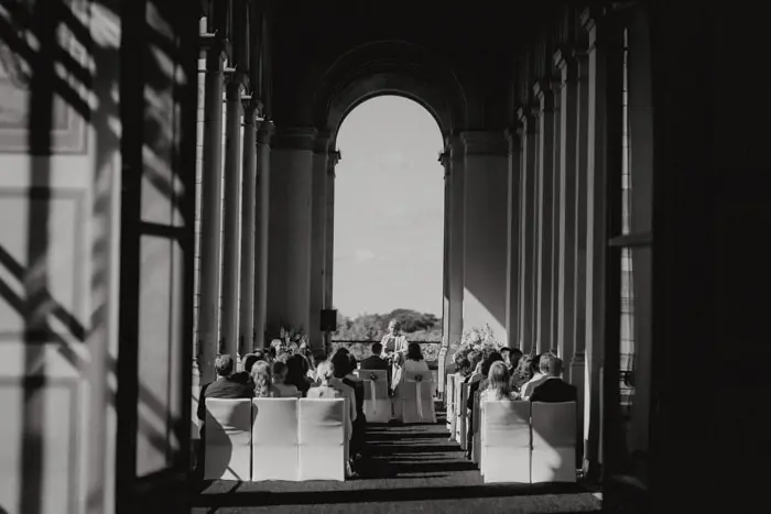 Hochzeitsfotograf Bayern Hochzeitszeremonie im Freien, Menschen sitzen zwischen Säulen Dokumentarische Editorial Hochzeitsfotografie