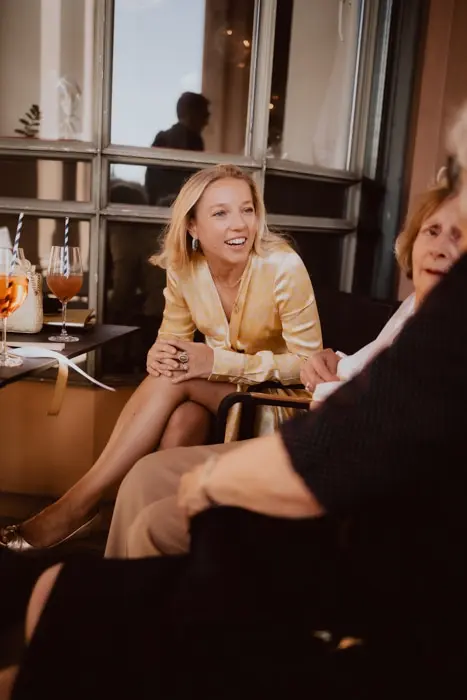 Hochzeitsfotograf Bayern Junge Frau in eleganter Kleidung sitzt und lächelt in gesellschaftlicher Runde Dokumentarische Editorial Hochzeitsfotografie