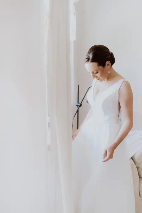 Hochzeitsfotograf Bayern Braut im eleganten weißen Kleid, steht nachdenklich vor hellem Hintergrund Dokumentarische Editorial Hochzeitsfotografie