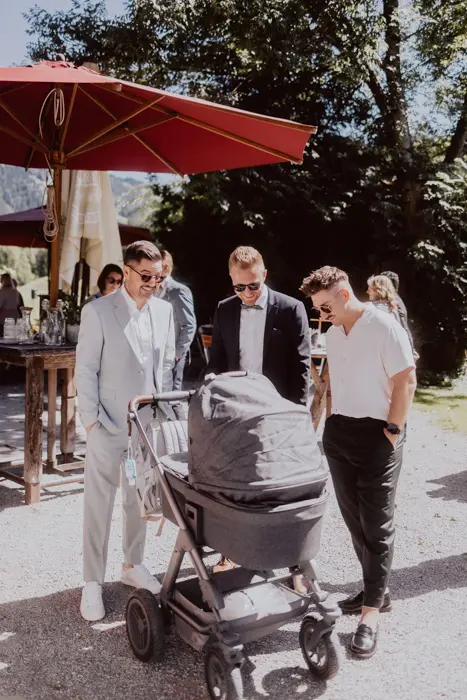 Hochzeitsfotograf Bayern Drei Männer betrachten Kinderwagen im Freien bei sonnigem Wetter Dokumentarische Editorial Hochzeitsfotografie