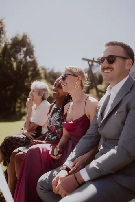 Hochzeitsfotograf Bayern Lächelnde Hochzeitsgäste im Freien bei sonnigem Wetter, lässig und festlich gekleidet Dokumentarische Editorial Hochzeitsfotografie