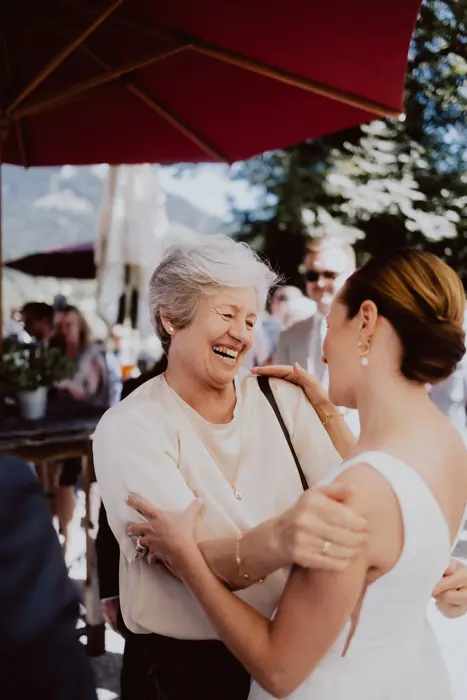 Hochzeitsfotograf Bayern Ältere Frau umarmt lachende Braut bei einer Outdoor-Hochzeitsfeier Dokumentarische Editorial Hochzeitsfotografie
