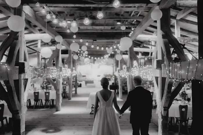 Hochzeitsfotograf Bayern Hochzeitspaar geht in festlich beleuchtetem Scheunenhochzeitssaal Dokumentarische Editorial Hochzeitsfotografie