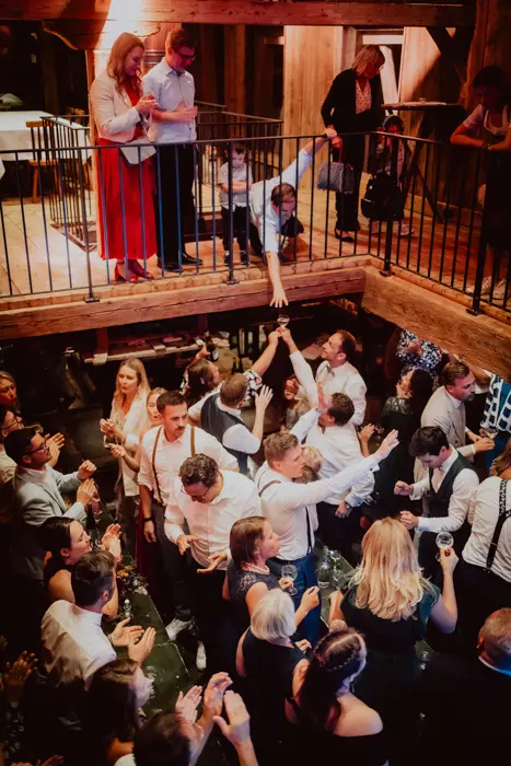 Hochzeitsfotograf Bayern Festliche Menschenmenge auf einer zweistöckigen Party mit Holzbalkonen Dokumentarische Editorial Hochzeitsfotografie