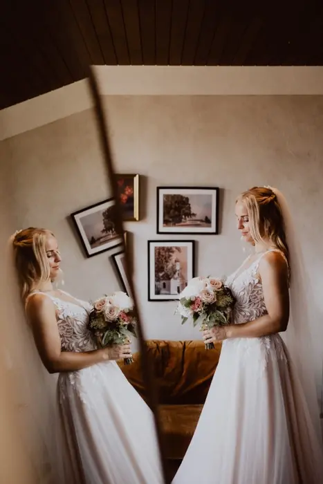 A bride in a white wedding dress holding a bouquet stands beside a mirror, creating a reflection. Framed photos hang on the wall behind her.