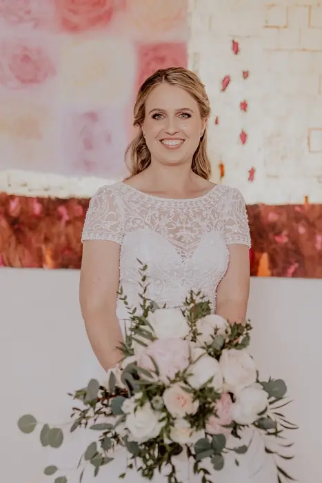 Hochzeitsfotograf Bayern Lächelnde Braut in weißem Kleid mit Blumenstrauß vor farbigem Hintergrund Dokumentarische Editorial Hochzeitsfotografie