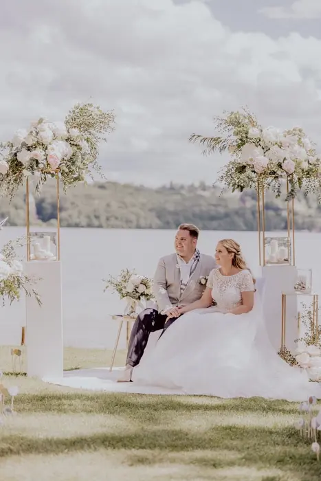 Hochzeitsfotograf Bayern Brautpaar bei einer Hochzeit am See mit blumengeschmücktem Hintergrund Dokumentarische Editorial Hochzeitsfotografie