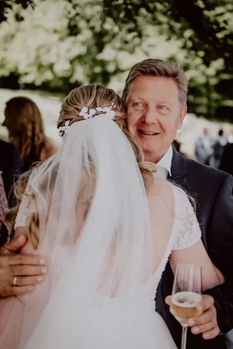 Hochzeitsfotograf Bayern Vater umarmt Braut bei Hochzeit im Freien im Sonnenschein Dokumentarische Editorial Hochzeitsfotografie