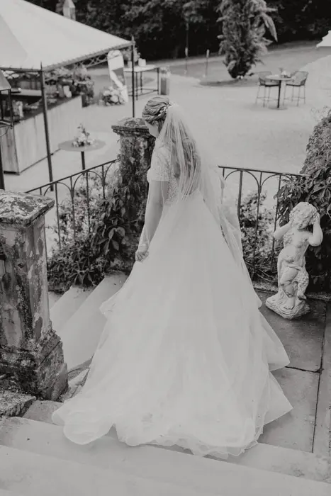 Hochzeitsfotograf Bayern Braut in Kleid mit Schleier geht Treppe im Freien hinunter Dokumentarische Editorial Hochzeitsfotografie