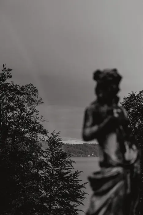 Hochzeitsfotograf Bayern Schwarzweiß-Foto einer Statue vor einem bewaldeten See Dokumentarische Editorial Hochzeitsfotografie