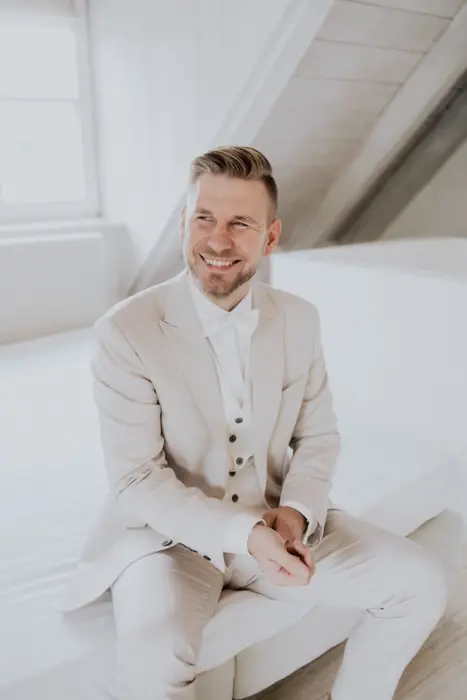 Hochzeitsfotograf Bayern Lächelnder Mann im eleganten weißen Anzug in einem hellen Raum Dokumentarische Editorial Hochzeitsfotografie