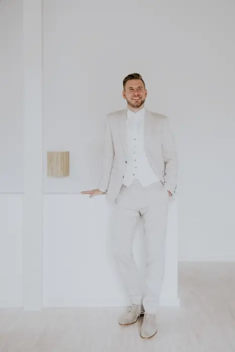 Hochzeitsfotograf Bayern Mann in weißem Anzug lehnt lächelnd an einer Wand Dokumentarische Editorial Hochzeitsfotografie