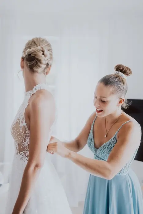 Hochzeitsfotograf Bayern Braut im weißen Kleid wird von der Trauzeugin angezogen Dokumentarische Editorial Hochzeitsfotografie