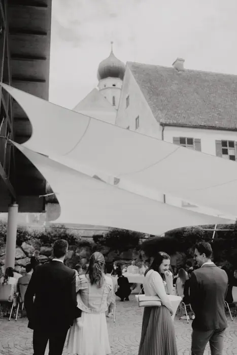 Hochzeitsfotograf Bayern Schwarzweißfoto einer Hochzeit mit Gästen im Freien und Kirche im Hintergrund Dokumentarische Editorial Hochzeitsfotografie