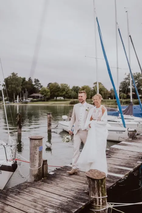 Hochzeitsfotograf Bayern Brautpaar spaziert auf einem Holzsteg am Seeufer bei Segelbooten Dokumentarische Editorial Hochzeitsfotografie