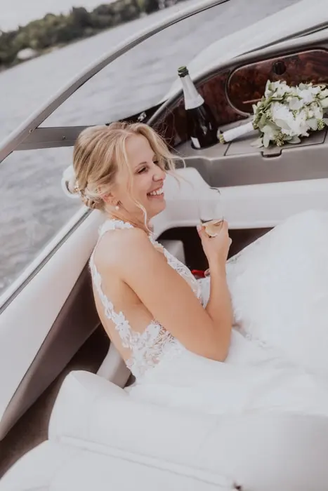 Hochzeitsfotograf Bayern Lächelnde Braut in weißem Kleid auf einem Boot mit Champagner Dokumentarische Editorial Hochzeitsfotografie