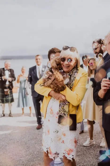 Hochzeitsfotograf Bayern Ältere Frau in gelber Jacke umarmt kleinen Hund bei einer Hochzeit Dokumentarische Editorial Hochzeitsfotografie