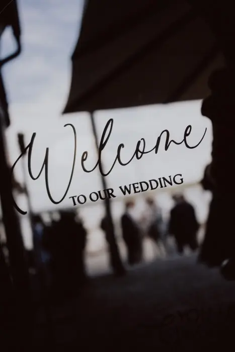 Hochzeitsfotograf Bayern Willkommensschild zur Hochzeit mit unscharfen Personen im Hintergrund Dokumentarische Editorial Hochzeitsfotografie