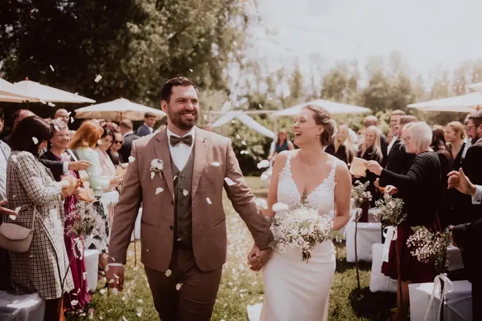 Hochzeitsfotograf Bayern Hochzeitspaar im Freien, lächelnd und händchenhaltend bei Blumendekoration Dokumentarische Editorial Hochzeitsfotografie