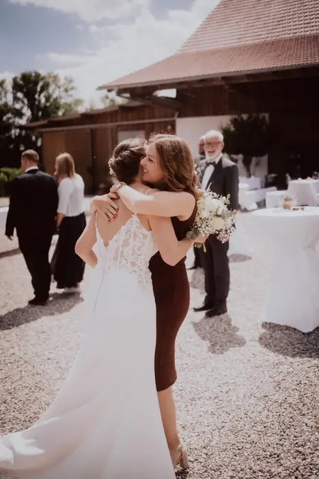 Hochzeitsfotograf Bayern Braut umarmt Freundin auf sonniger Hochzeit im Freien Dokumentarische Editorial Hochzeitsfotografie
