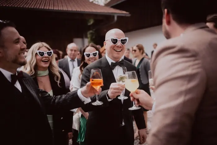 Hochzeitsfotograf Bayern Fröhliche Hochzeitsgäste mit herzförmigen Sonnenbrillen stoßen im Freien an Dokumentarische Editorial Hochzeitsfotografie