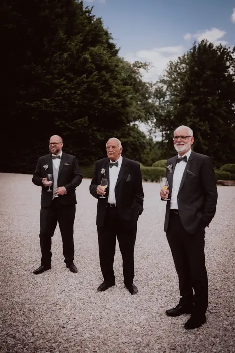 Hochzeitsfotograf Bayern Drei Männer in Abendgarderobe stehen auf einem Kiesweg im Freien Dokumentarische Editorial Hochzeitsfotografie