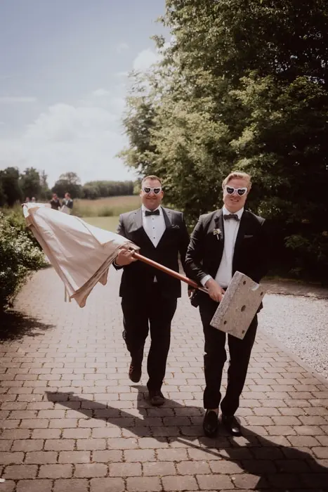 Hochzeitsfotograf Bayern Zwei Männer in Anzügen mit Requisit im Freien Dokumentarische Editorial Hochzeitsfotografie