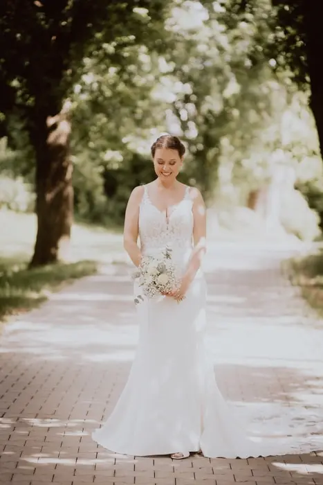 Hochzeitsfotograf Bayern Braut in weißem Kleid auf sonnigem Parkweg mit Bäumen Dokumentarische Editorial Hochzeitsfotografie