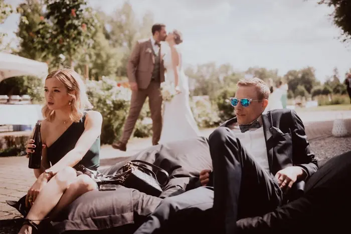 Hochzeitsfotograf Bayern Hochzeitspaar im Hintergrund, Gäste im Vordergrund auf Sitzsäcken im Freien Dokumentarische Editorial Hochzeitsfotografie