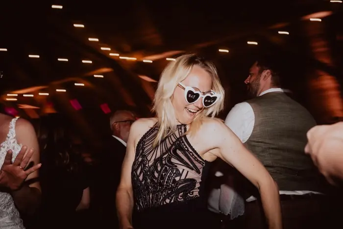 Hochzeitsfotograf Bayern Frau in elegantem Kleid tanzt auf einer Party mit Herz-Sonnenbrille Dokumentarische Editorial Hochzeitsfotografie