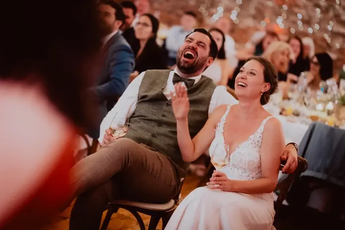 Hochzeitsfotograf Bayern Hochzeitspaar lacht während Empfangsitze auf Stühlen Dokumentarische Editorial Hochzeitsfotografie