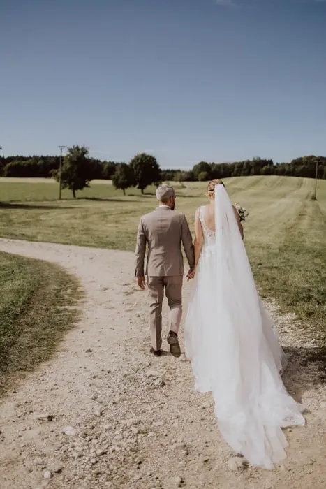 Hochzeitsfotograf Bayern Brautpaar spaziert Hand in Hand auf ländlichem Weg Dokumentarische Editorial Hochzeitsfotografie