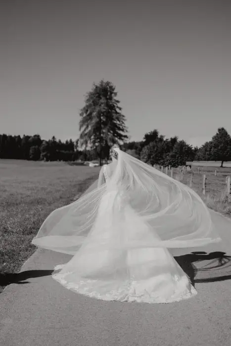 Hochzeitsfotograf Bayern Braut in fließendem Hochzeitskleid auf Landstraße mit Schleier im Wind Dokumentarische Editorial Hochzeitsfotografie