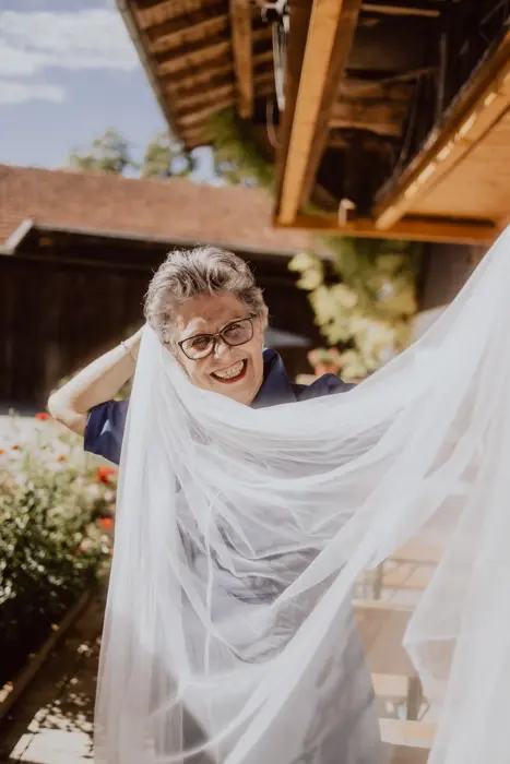 Hochzeitsfotograf Bayern Fröhliche Person mit Brille hält weißen Schleier im Garten Dokumentarische Editorial Hochzeitsfotografie