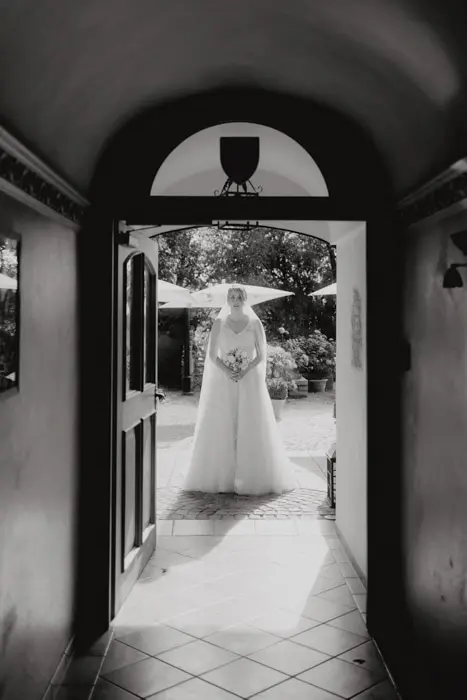 Hochzeitsfotograf Bayern Braut im weißen Kleid steht vor einer Tür im Garten Dokumentarische Editorial Hochzeitsfotografie