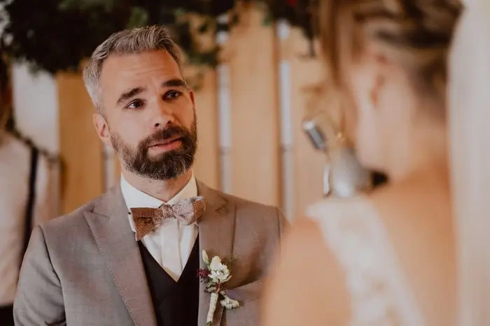 Hochzeitsfotograf Bayern Bräutigam im grauen Anzug blickt Braut während Hochzeitszeremonie an Dokumentarische Editorial Hochzeitsfotografie