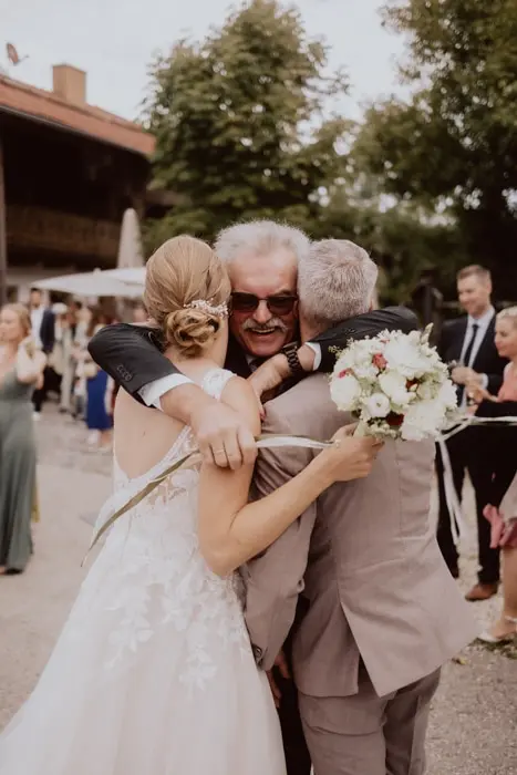 Hochzeitsfotograf Bayern Brautpaar umarmt älteren Gast auf Hochzeitsempfang im Freien Dokumentarische Editorial Hochzeitsfotografie