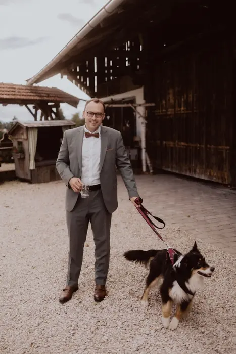 Hochzeitsfotograf Bayern Mann im Anzug führt Hund an der Leine vor einer Scheune Dokumentarische Editorial Hochzeitsfotografie