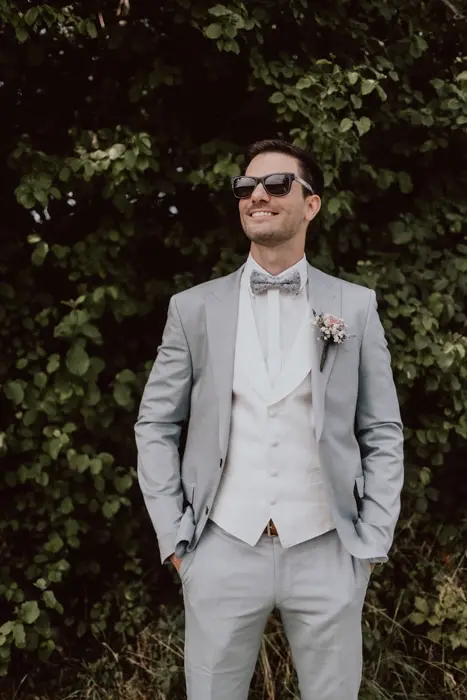 Hochzeitsfotograf Bayern Mann im grauen Anzug mit Sonnenbrille vor Laubwand Dokumentarische Editorial Hochzeitsfotografie